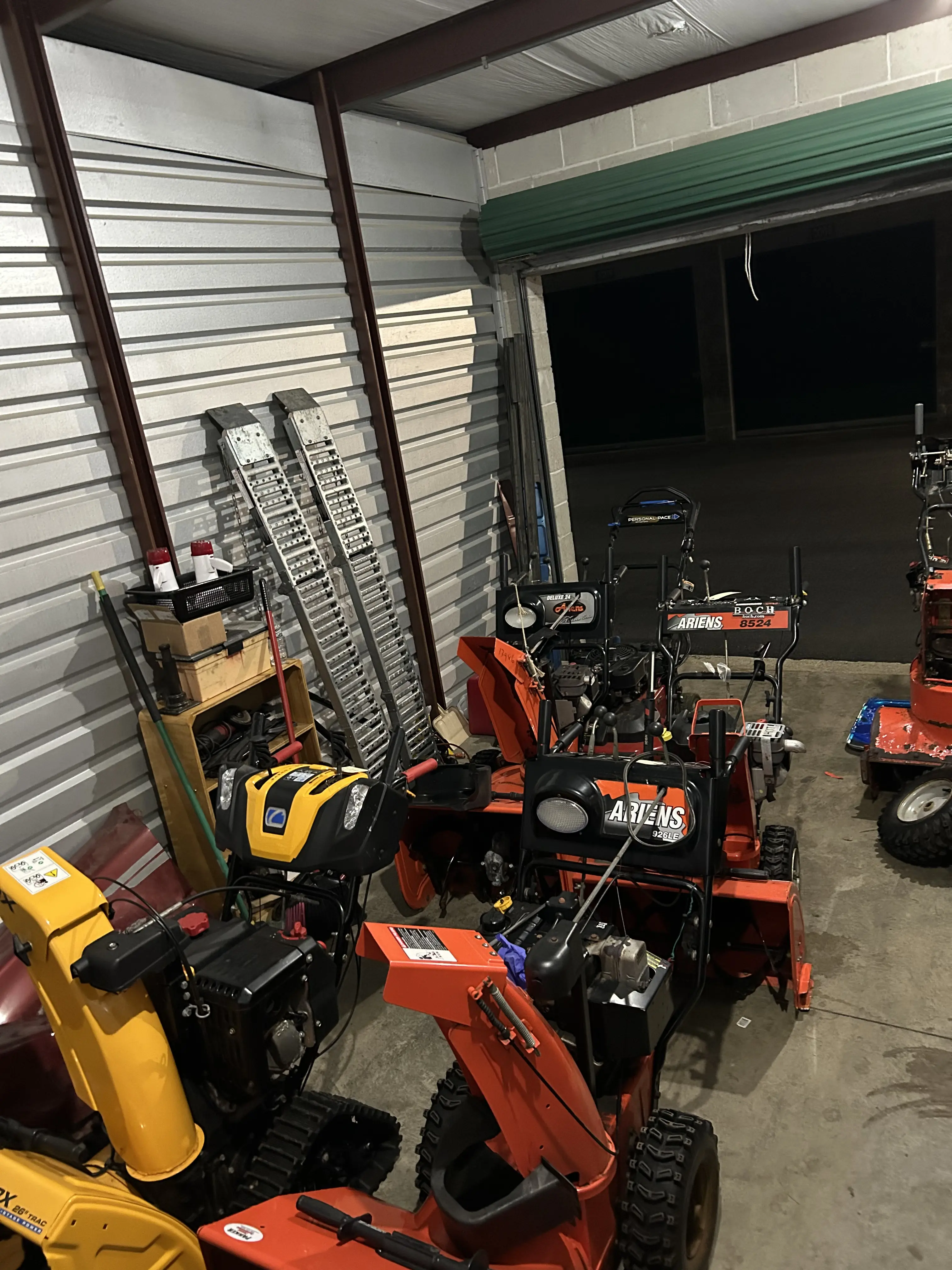 Inside the shop bay: a row of snowblowers and equipment lined up along a corrugated metal wall, ramps standing ready.