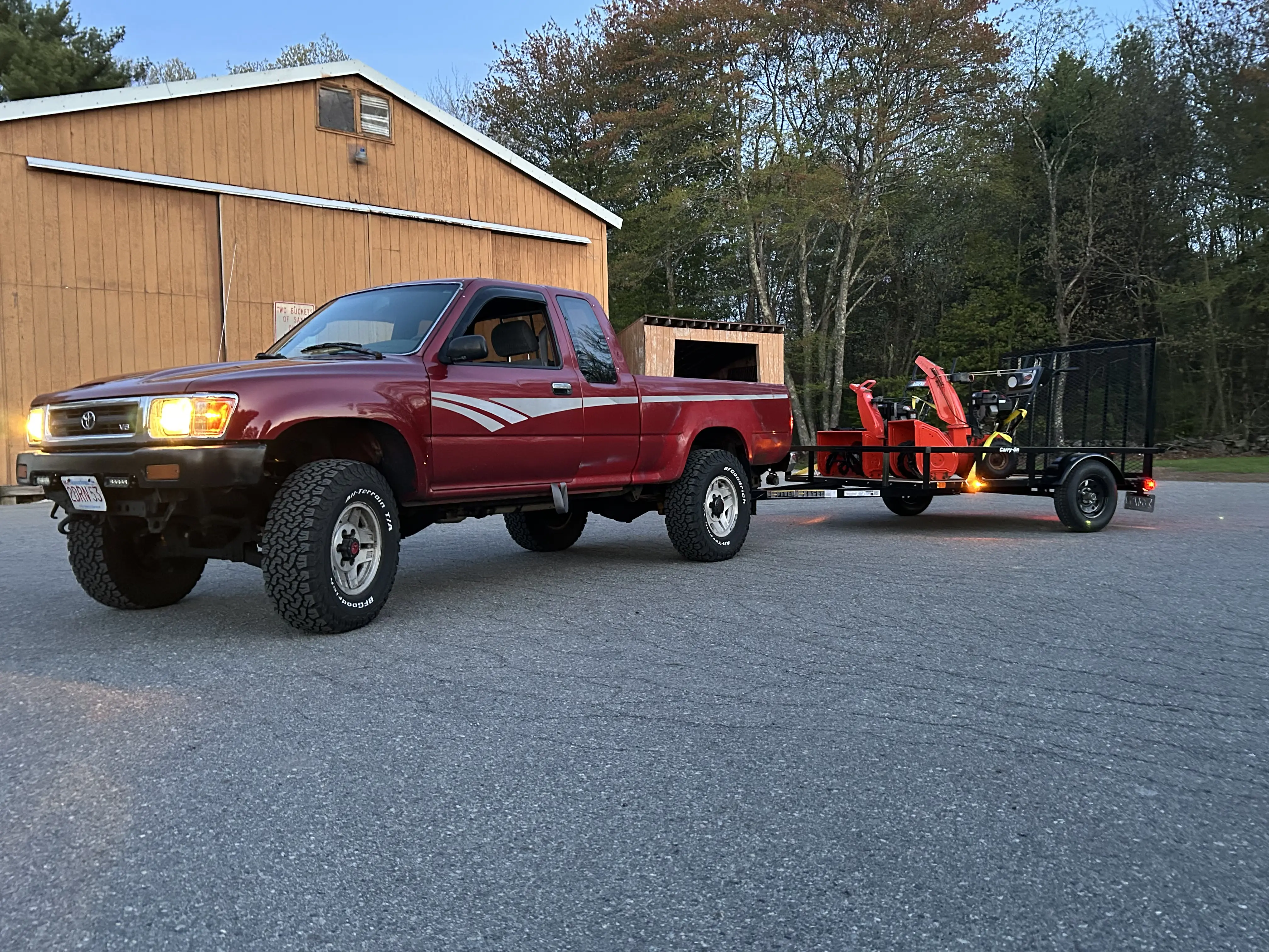 A red Toyota pickup at golden hour towing a trailer with a snowblower aboard, parked by a barn.